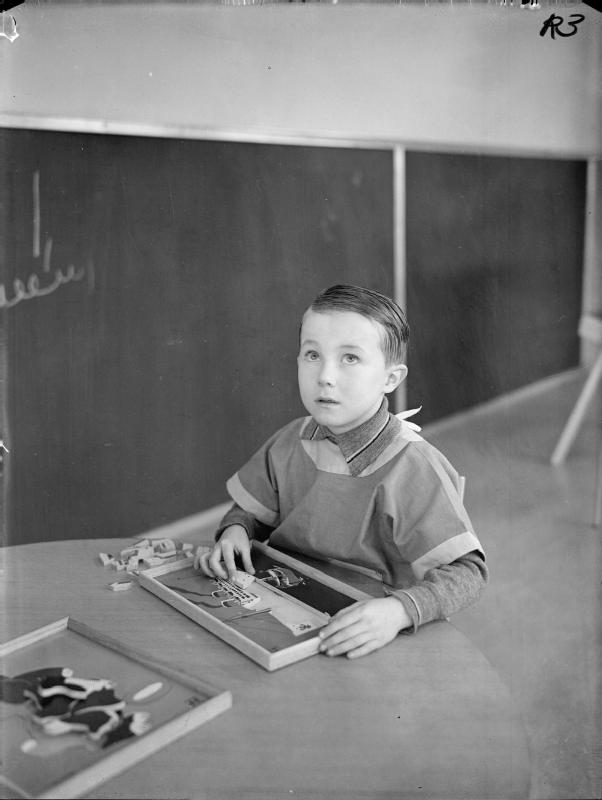 A young boy in painting overalls plays with a wooden jigsaw puzzle at Park Lane Municipal Day Nursery in Tottenham, London, 1940. The black-and-white wartime photograph shows the child concentrating on fitting pieces together at a small table.