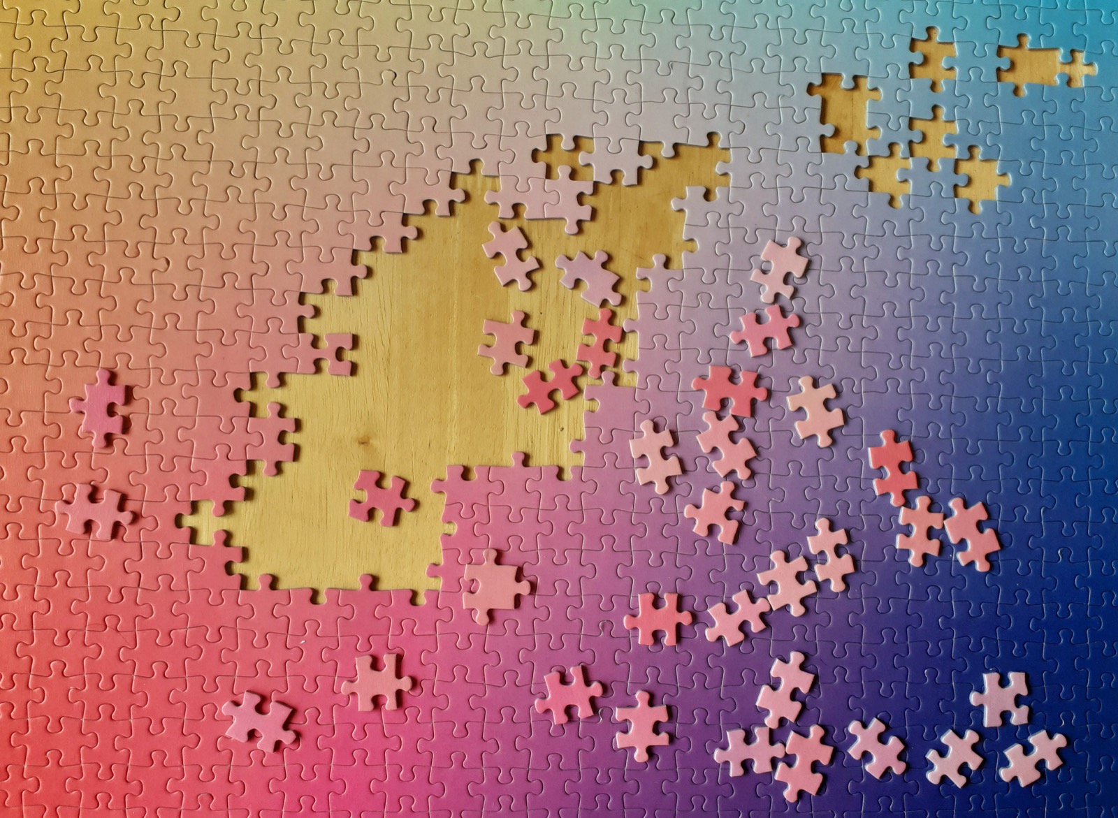 An incomplete 1,000-piece gradient jigsaw puzzle on a wooden table, its colors shifting from pink through purple to deep blue. A gap in the center reveals the table underneath, with scattered unplaced pieces nearby.