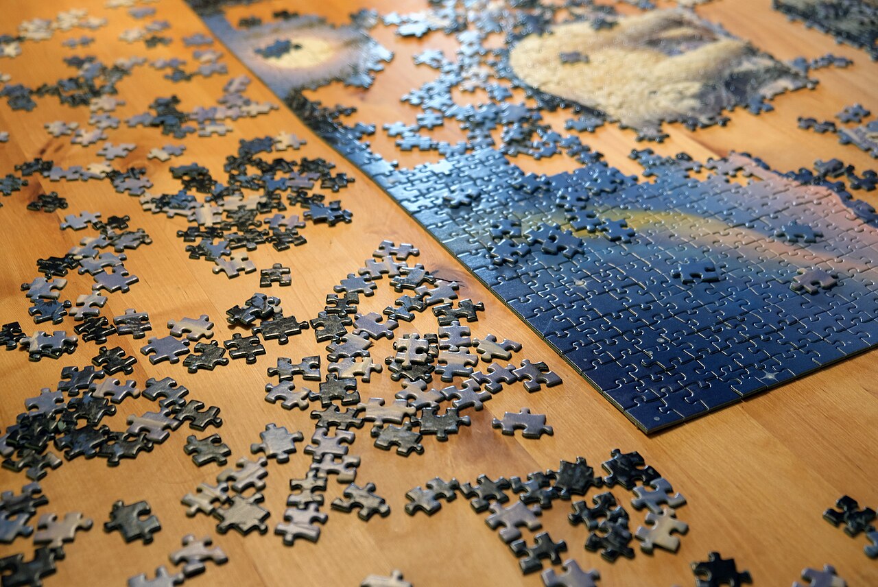 A jigsaw puzzle in progress on a wooden table, seen from above. Scattered pieces surround a partially assembled section, with a reference image visible nearby. The quiet, solitary scene captures the private nature of puzzling that the article describes.