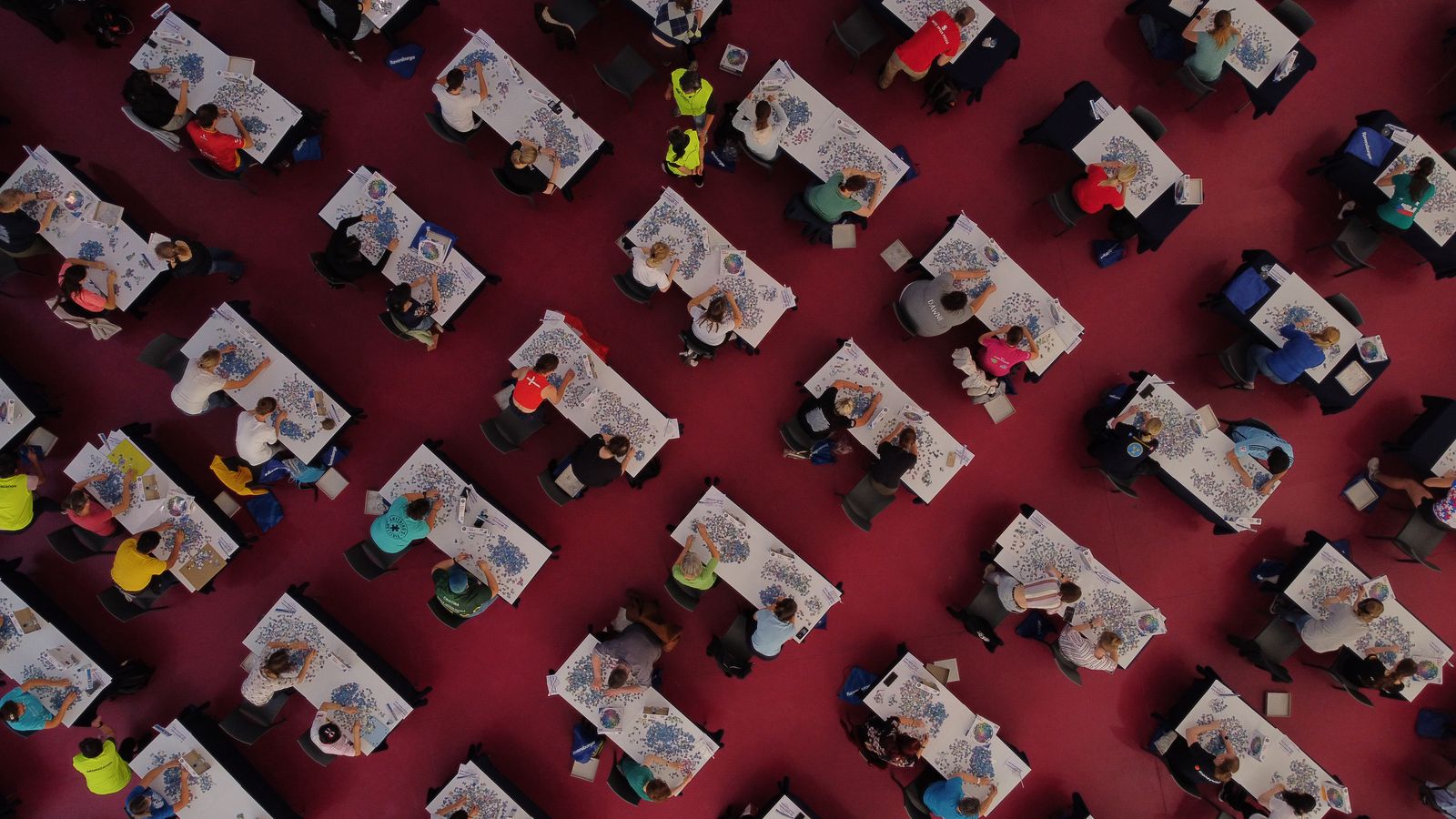 Aerial drone photograph of the 2023 World Jigsaw Puzzle Championship individual final in Valladolid, Spain. Hundreds of competitors sit at rows of tables in a large arena, each racing to complete a jigsaw puzzle as fast as possible.